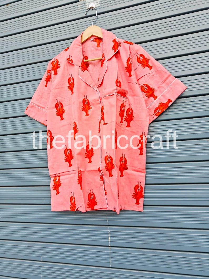 May include: Pink pajama set with a red lobster print. The short-sleeved top has a notched collar, a chest pocket, and white trim. The fabric is hanging on a wooden hanger.