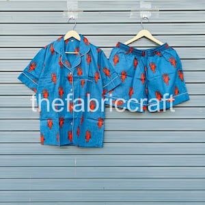 May include: Blue pajama set with a red lobster pattern. The set includes a short-sleeved button-down shirt and matching shorts. The shirt and shorts have white trim and are hanging on wooden hangers.
