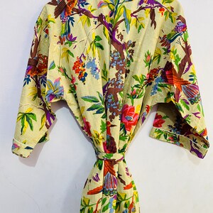 以下が含まれることがあります： カラフルな鳥と花が描かれた花柄のベージュの綿のローブ。ローブにはリボンベルトと長袖が付いています。