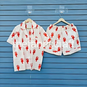 May include: A white pajama set with a red lobster print. The set includes a short-sleeved button-down shirt and matching shorts. The shirt has a collar and a chest pocket. The shorts have an elastic waistband.