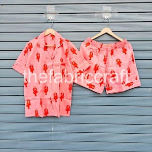 May include: A pink pajama set with a red lobster pattern. The set includes a button-down shirt with a chest pocket and shorts. The fabric is lightweight and the set is hanging on wooden hangers.