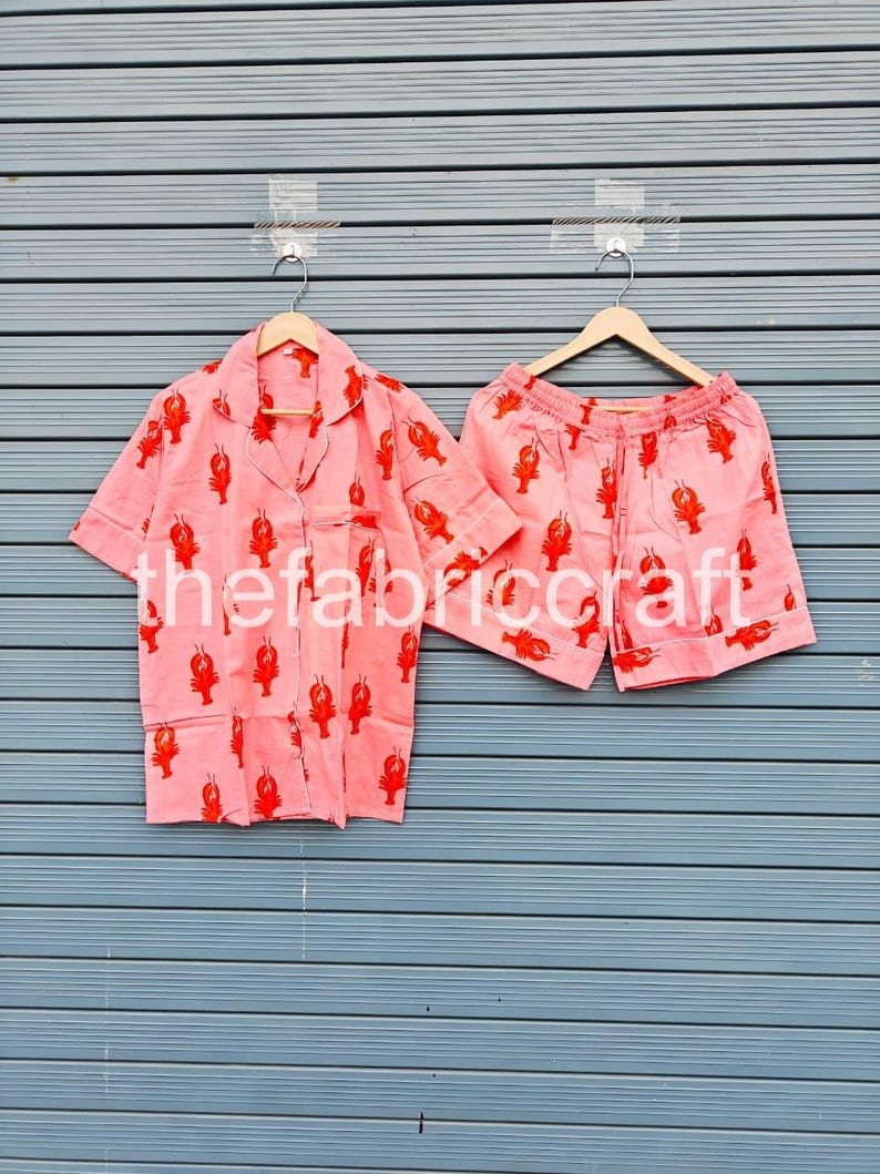 May include: A pink pajama set with a red lobster pattern. The set includes a button-down shirt with a chest pocket and shorts. The fabric is lightweight and the set is hanging on wooden hangers.