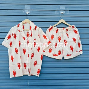 May include: A white pajama set with a red lobster pattern. The set includes a short-sleeved button-down shirt and matching shorts. The fabric is lightweight, and the design is repeated across both pieces. The set is hanging on a wooden hanger.