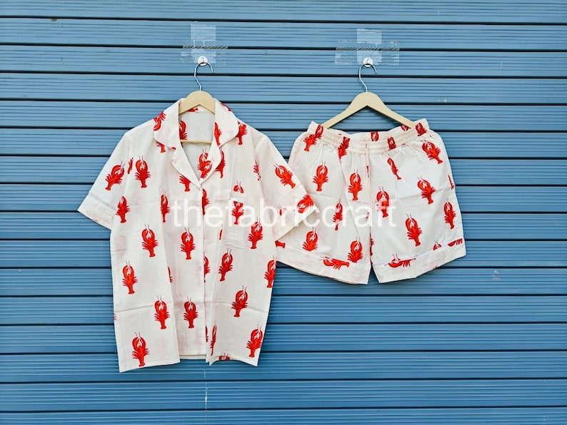 May include: A white pajama set with a red lobster pattern. The set includes a short-sleeved button-down shirt and matching shorts. The fabric is lightweight, and the design is repeated across both pieces. The set is hanging on a wooden hanger.