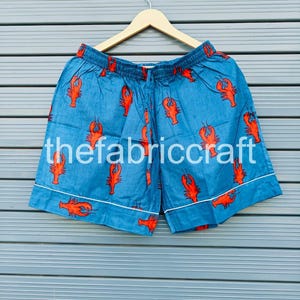 May include: Blue shorts with a red lobster pattern and white trim. The text "thefabriccraft" is printed on the shorts. The shorts are hanging on a wooden hanger against a blue background.