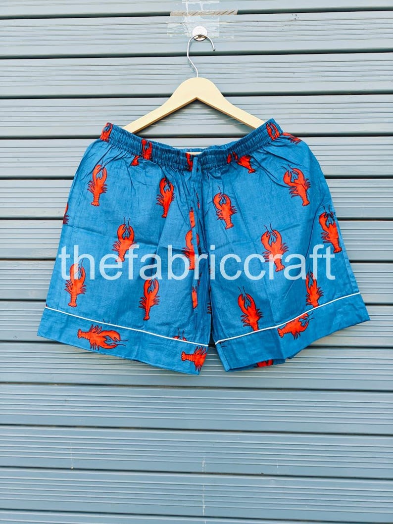 May include: Blue shorts with a red lobster pattern and white trim. The text "thefabriccraft" is printed on the shorts. The shorts are hanging on a wooden hanger against a blue background.