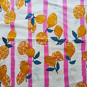 May include: A white fabric featuring a repeating pattern of yellow lemons and orange citrus slices with dark teal leaves, arranged over vertical pink stripes. The text thefabricraft is printed in white across the center of the material.