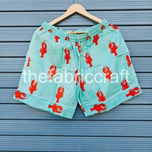May include: Light blue shorts with a repeating pattern of red lobsters. The shorts have a white trim and a drawstring waistband. The text "thefabriccraft" is printed on the shorts.