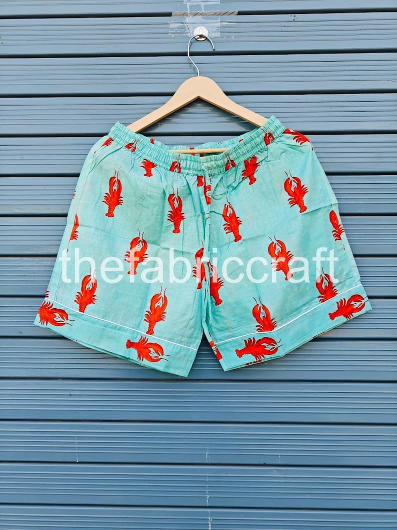 May include: Light blue shorts with a repeating pattern of red lobsters. The shorts have a white trim and a drawstring waistband. The text "thefabriccraft" is printed on the shorts.