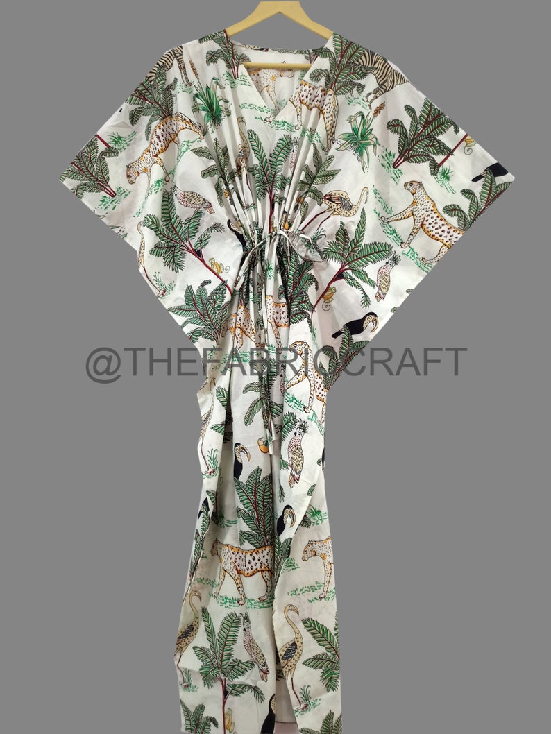 Op de afbeelding: Witte katoenen kaftan met een groene, zwarte en bruine dierenprint. De print bevat luipaarden, toekans en palmbomen. De kaftan heeft een V-hals en een tailleband.