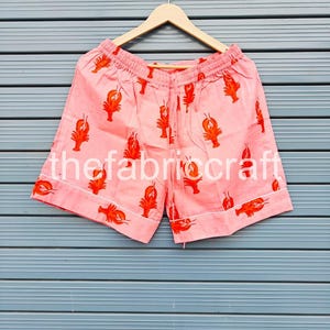 May include: Pink shorts with a repeating pattern of orange lobsters. The shorts have a drawstring waist and white trim. The text "thefabricraft" is printed on the shorts. The shorts are hanging on a wooden hanger.