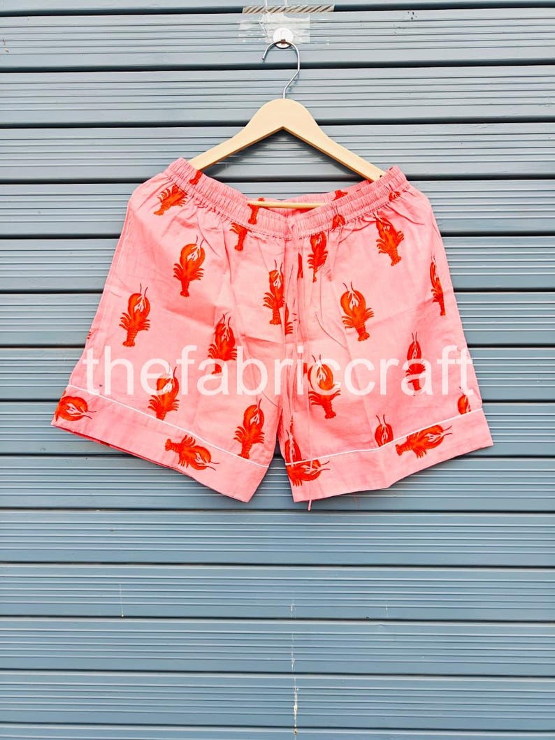 May include: Pink shorts with a repeating pattern of orange lobsters. The shorts have a drawstring waist and white trim. The text "thefabricraft" is printed on the shorts. The shorts are hanging on a wooden hanger.