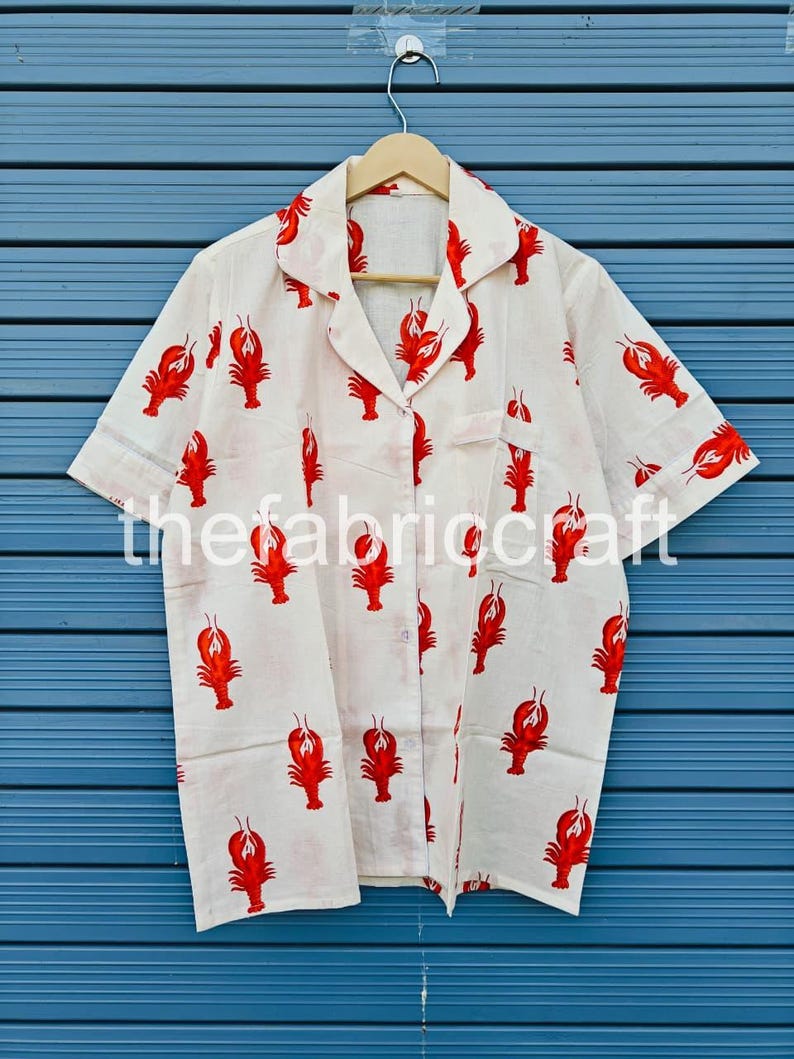 May include: White button-down shirt with short sleeves, featuring a repeating pattern of red lobsters. The shirt has a collared neckline and a breast pocket. The text "thefabriccraft" is printed on the shirt.