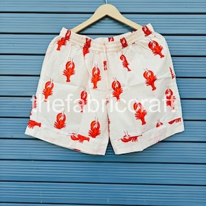 May include: Cream-colored shorts with a repeating pattern of red lobsters. The shorts have an elastic waistband and are hanging on a wooden hanger against a blue background. The shorts are made of a lightweight fabric.