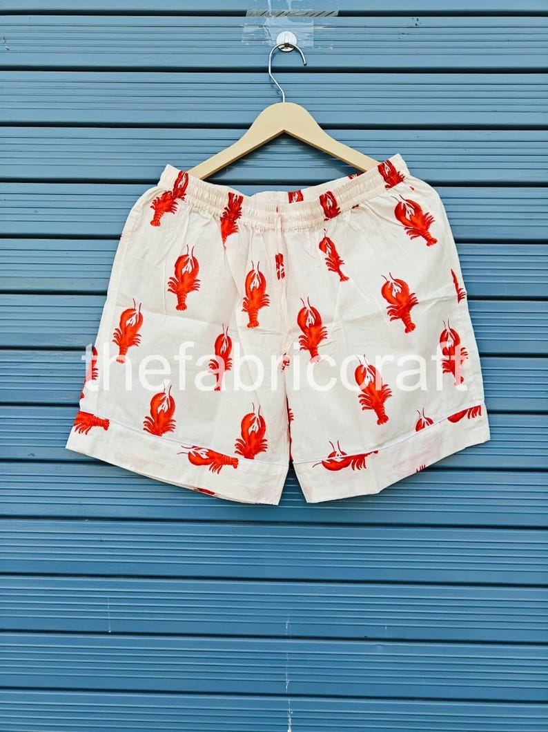 May include: Cream-colored shorts with a repeating pattern of red lobsters. The shorts have an elastic waistband and are hanging on a wooden hanger against a blue background. The shorts are made of a lightweight fabric.