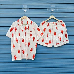 May include: A white pajama set with a red lobster print. The set includes a short-sleeved button-down shirt and matching shorts. The shirt has a collar and a chest pocket. The shorts have an elastic waistband.