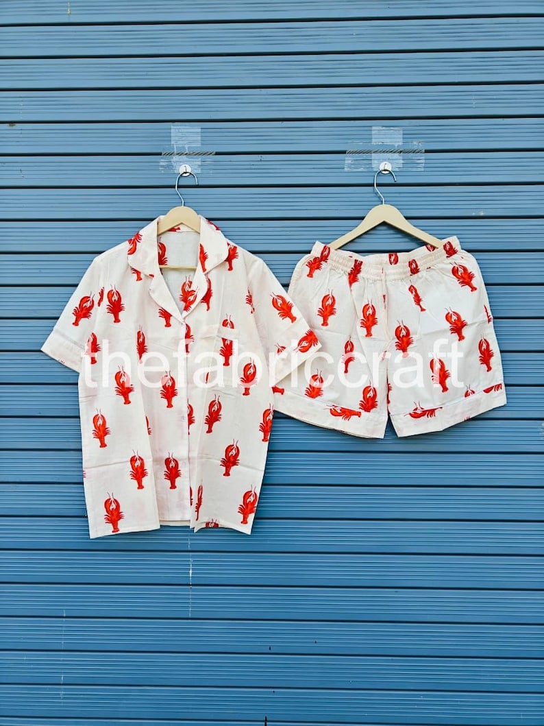 May include: A white pajama set with a red lobster print. The set includes a short-sleeved button-down shirt and matching shorts. The shirt has a collar and a chest pocket. The shorts have an elastic waistband.