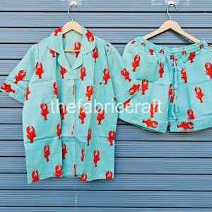 May include: A light blue pajama set featuring a red lobster print. The set includes a short-sleeved button-up shirt and matching shorts. The fabric is lightweight and the set is hanging on a wooden hanger.