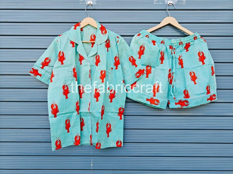 May include: A light blue pajama set featuring a red lobster print. The set includes a short-sleeved button-up shirt and matching shorts. The fabric is lightweight and the set is hanging on a wooden hanger.