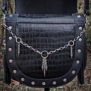 May include: Black crossbody bag with a faux crocodile skin texture. The bag features silver-toned studs, a chain detail with dangling spikes, and a tassel on each side. The bag has a shoulder strap.