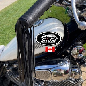 May include: Black leather motorcycle handlebar grips with fringe detail. The grips are attached to a motorcycle with a white gas tank. The logo for Bristol is visible on the gas tank.