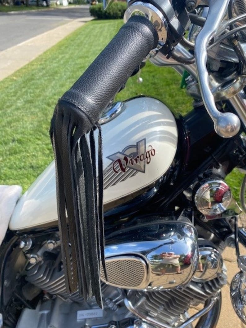 May include: Close-up of a motorcycle handlebar with a black leather grip and fringe. The motorcycle has a cream-colored gas tank with the word "Virago" in red script. Chrome accents and a shiny engine are also visible.