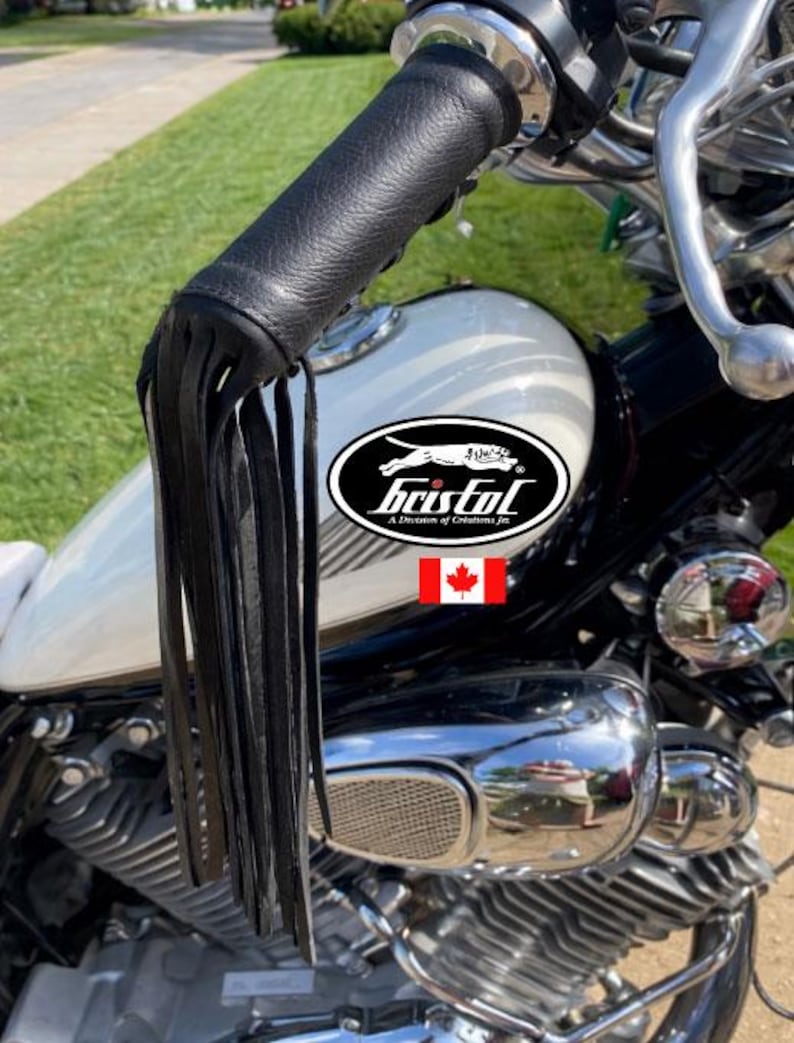 May include: Black leather motorcycle handlebar grips with fringe detail. The grips are attached to a motorcycle with a white gas tank. The logo for Bristol is visible on the gas tank.