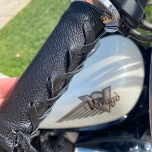 May include: Black leather motorcycle handlebar grip with fringe detail. The grip is textured and laced, with a chrome accent and a portion of the motorcycle's white fuel tank visible.