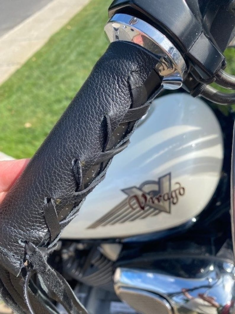 May include: Black leather motorcycle handlebar grip with fringe detail. The grip is textured and laced, with a chrome accent and a portion of the motorcycle's white fuel tank visible.