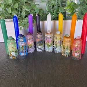 May include: Ten small glass bottles filled with crystals, herbs, and other natural elements. The bottles are arranged in a row on a wooden surface, with a candle of each color of the rainbow behind them.