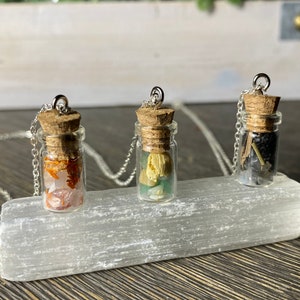 May include: Three small glass bottles with cork stoppers and silver chains, each filled with different colored crystals and stones. The bottles are displayed on a white selenite slab.