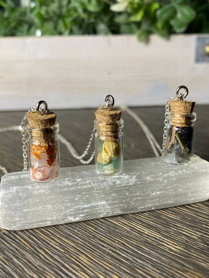 May include: Three small glass bottles with cork stoppers and silver chains, each filled with different colored crystals and stones. The bottles are displayed on a white selenite slab.