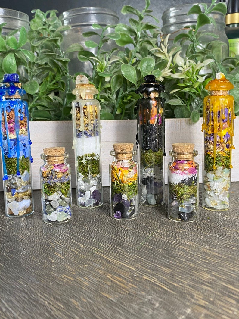 May include: Seven small glass bottles filled with various dried herbs, flowers, and crystals. The bottles are sealed with cork stoppers and have a layer of green moss at the bottom. The bottles are arranged in a row on a wooden surface.