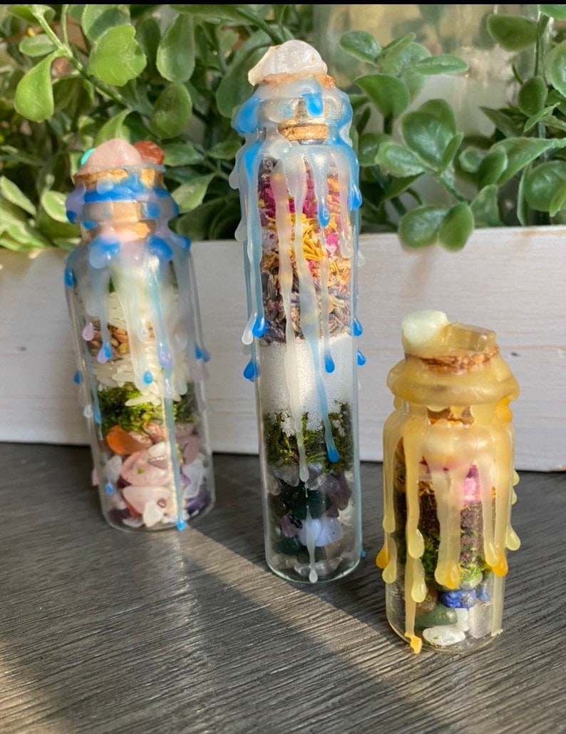 May include: Three glass bottles filled with various crystals, herbs, and dried flowers, sealed with cork stoppers and topped with a layer of wax. The bottles are decorated with blue, yellow, and white wax drippings.