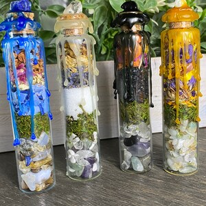 May include: Four small glass bottles filled with various crystals, herbs, and moss. Each bottle is topped with a different colored wax seal, including blue, white, black, and yellow.