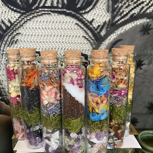 May include: Eight small glass bottles with cork stoppers filled with dried flowers, moss, and crystals. The bottles are arranged in a row on a wooden surface.