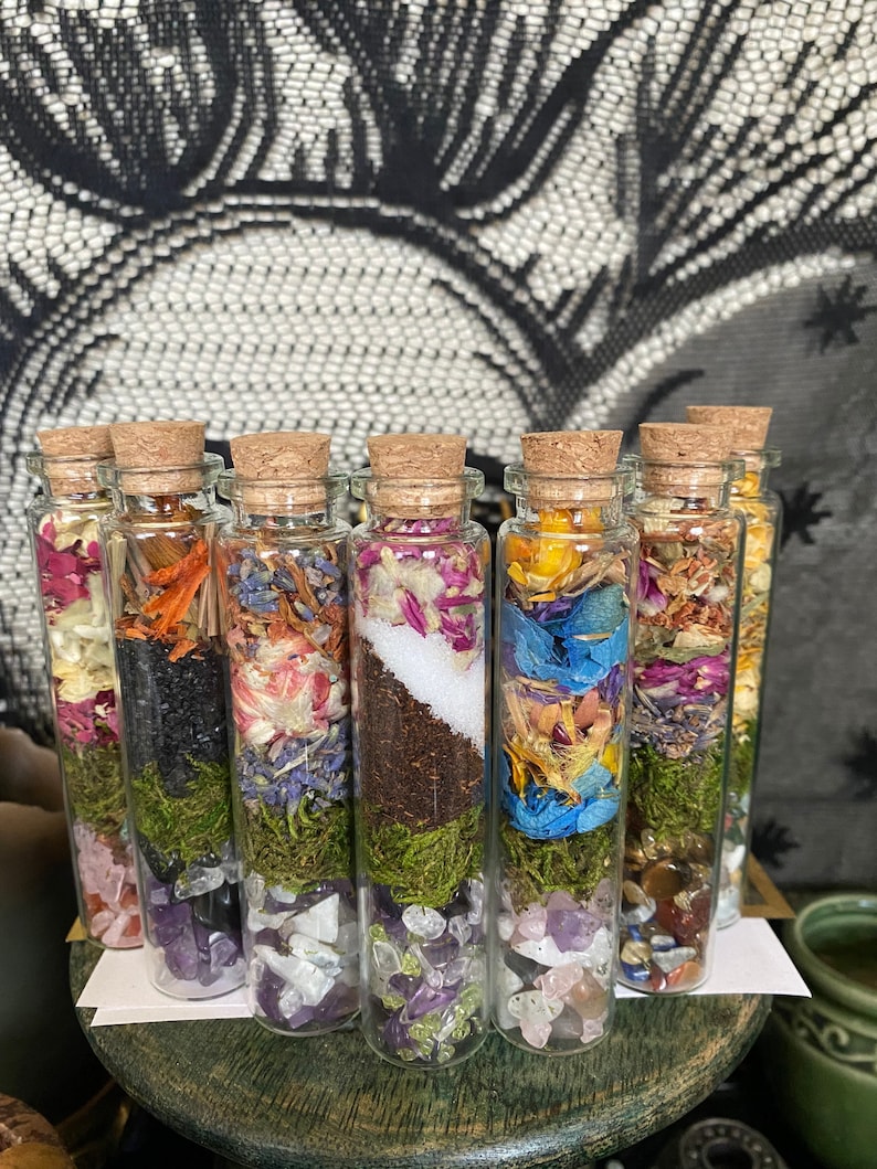 May include: Eight small glass bottles with cork stoppers filled with dried flowers, moss, and crystals. The bottles are arranged in a row on a wooden surface.