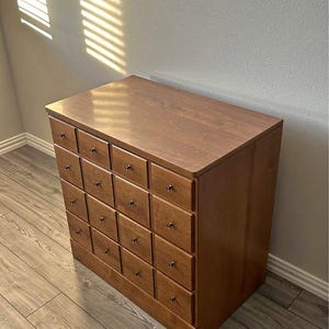 May include: A wooden chest of drawers with a warm brown finish. The chest has multiple small drawers with dark knobs. The top surface is smooth and the piece is set on a wooden floor.