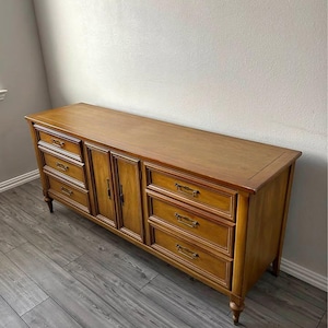 May include: A wooden dresser with a warm, honey-toned finish. It features multiple drawers and cabinet doors with brass-toned hardware. The dresser has a rectangular shape and stands on short, decorative legs.