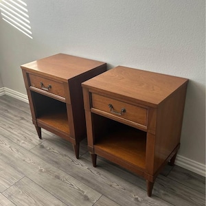 May include: Two matching wooden nightstands with a warm brown finish. Each nightstand features a drawer with a dark metal handle and an open shelf below. The nightstands have tapered legs and a classic design.