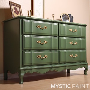 May include: A six-drawer dresser painted in a deep green hue. The dresser features ornate gold-colored drawer pulls and a scalloped base. The top of the dresser has decorative items, including a framed picture and a small mirror.