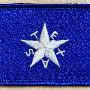 Puede incluir: Un parche rectangular azul con una estrella blanca en el centro, bordada con la palabra "TEXAS". El parche tiene un borde cosido. La estrella tiene las letras T, E, X, A y S bordadas.