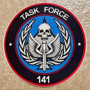 Könnte beinhalten: Runder Aufnäher mit dem Text "TASK FORCE" in Weiß, der ein zentrales Emblem umgibt. Das Emblem zeigt einen Totenkopf, Flügel und ein Schwert, mit der Nummer "141" unten. Das Design hat einen blauen und schwarzen Hintergrund mit roten und weißen Rändern.