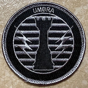 Puede incluir: Parche bordado circular en blanco y negro con la palabra "UMBRA" en la parte superior. El diseño presenta una torre estilizada en el centro, flanqueada por dos rayos. El parche tiene un borde detallado y cosido.