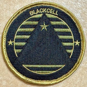 Peut inclure: Écusson brodé circulaire avec le mot "BLACKCELL" en arc de cercle en haut. Le patch présente un triangle noir au centre, entouré d'une bordure et d'accents dorés. Trois étoiles dorées sont placées autour du triangle.