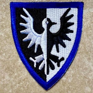 May include: Embroidered patch featuring a stylized bird design, split vertically into black and white halves. The patch has a shield shape with a blue border, suitable for jackets, bags, or other fabric items.