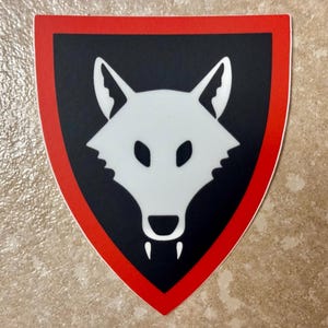 以下が含まれることがあります： 赤色の縁取りのある黒いシールド型のステッカー。白いオオカミの頭が描かれており、尖った耳、暗い目、牙が特徴です。これはパッチまたはエンブレムである可能性があります。