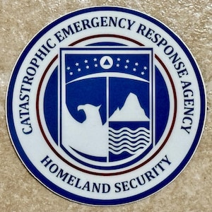 Könnte beinhalten: Ein runder Aufkleber mit dem Text "Catastrophic Emergency Response Agency" und "Homeland Security". Das Design zeigt ein blau-weißes Emblem mit einem Adler, Bergen und Wellen.