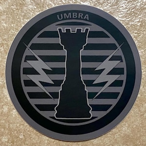 Puede incluir: Una pegatina redonda con un diseño en negro y gris. La palabra "UMBRA" está en la parte superior. El diseño presenta una pieza de ajedrez en el centro, con rayos a cada lado. El fondo tiene rayas horizontales.
