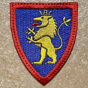 May include: Embroidered patch with a shield shape. The patch features a yellow lion with a crown, red tongue, and cross-shaped objects on a blue background. The shield is outlined with red stitching.